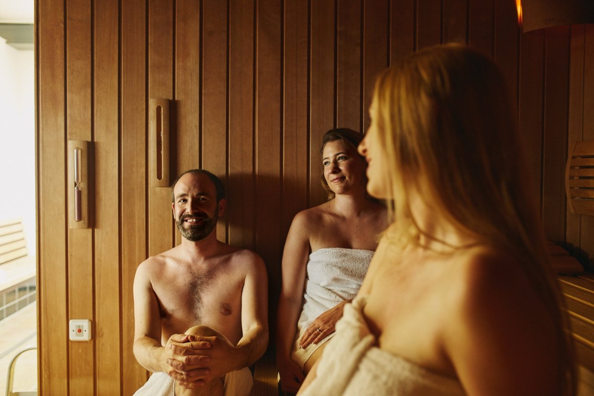 Im Vordergrund Frau die sich in der Sauna entspannt und eine weitere Frau, sowie ein Mann im Hintergrund. 