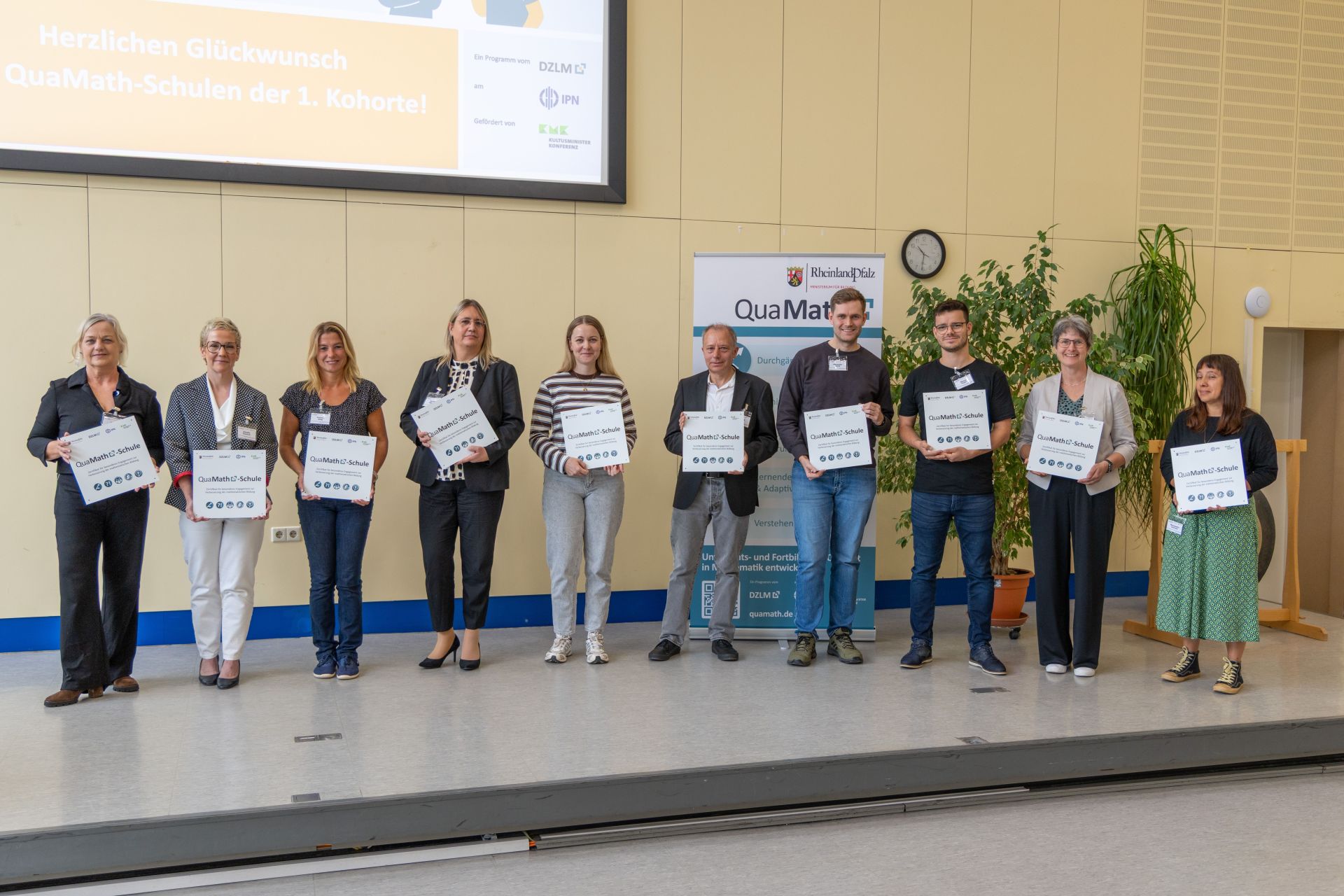 10 Menschen, die kleine Tafeln/Hinweisschilder  mit der Aufschrift QuaMath-Schule Richtung Kamera zeigen